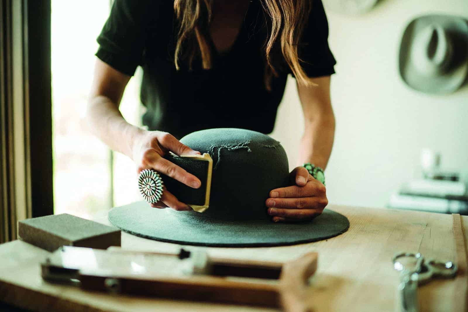In a rustic western story setting, a woman deftly crafts a hat on a wooden table.