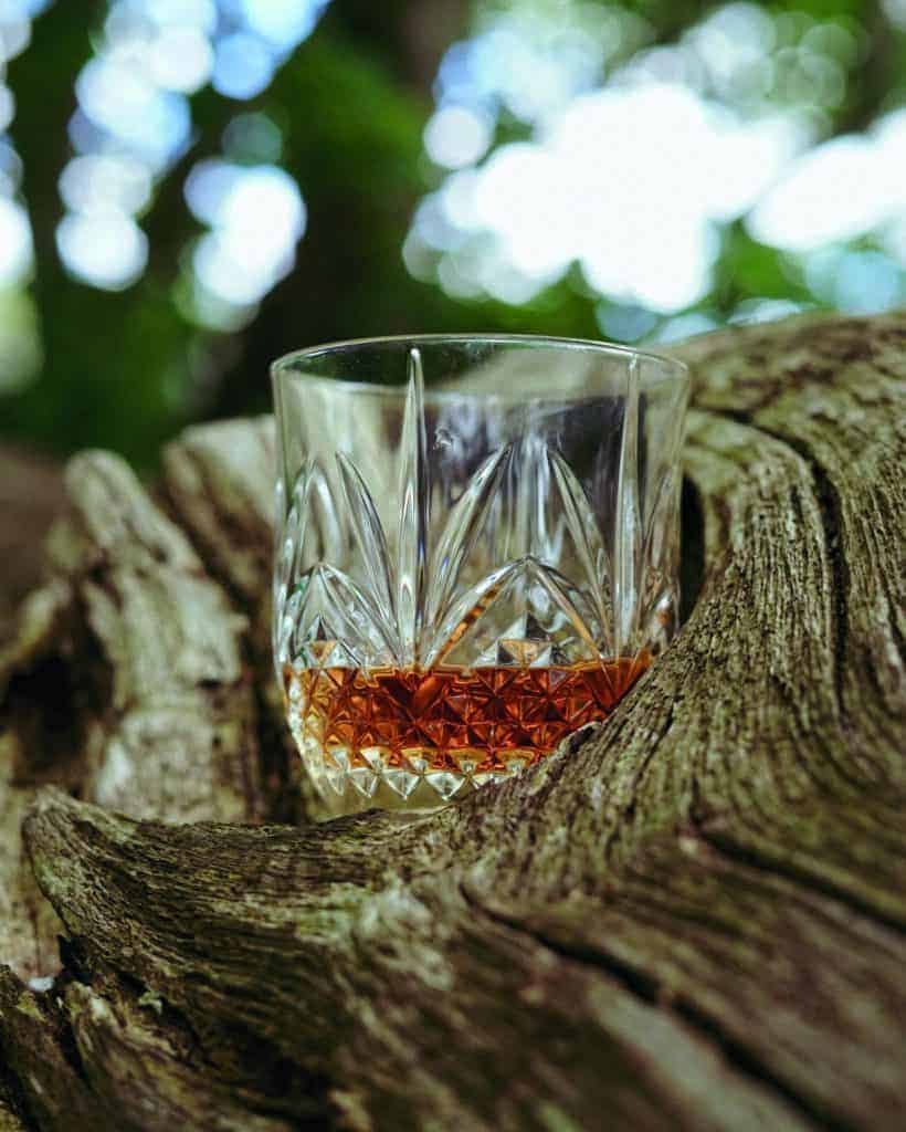A glass of whisky sitting on a tree trunk, next to a barrel.