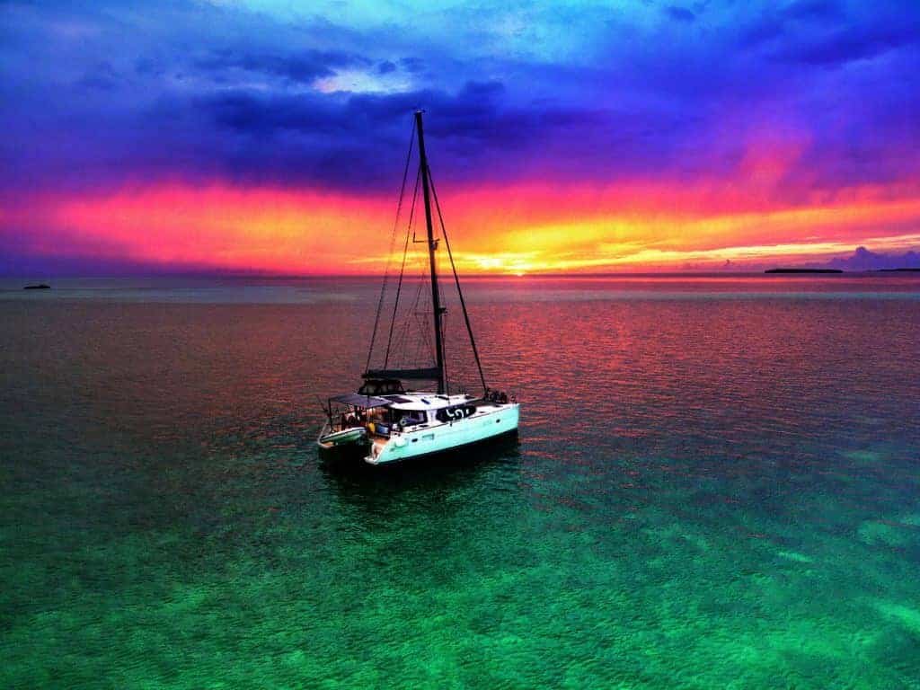 A catamaran sails away in the ocean at sunset.
