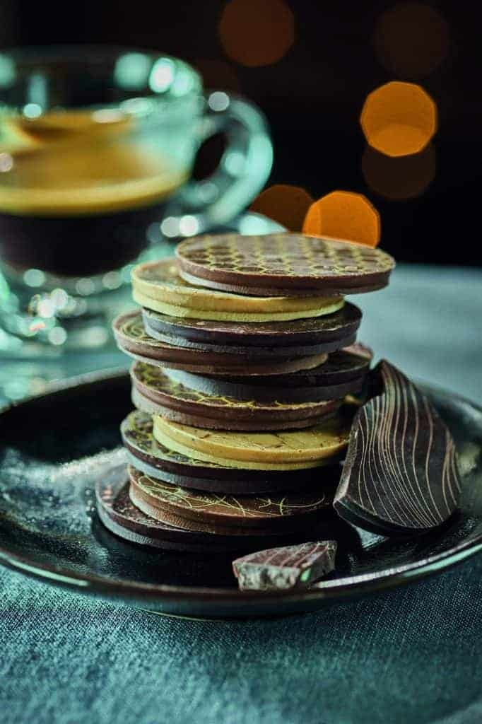 A stack of chocolates on a plate next to a cup of coffee.
